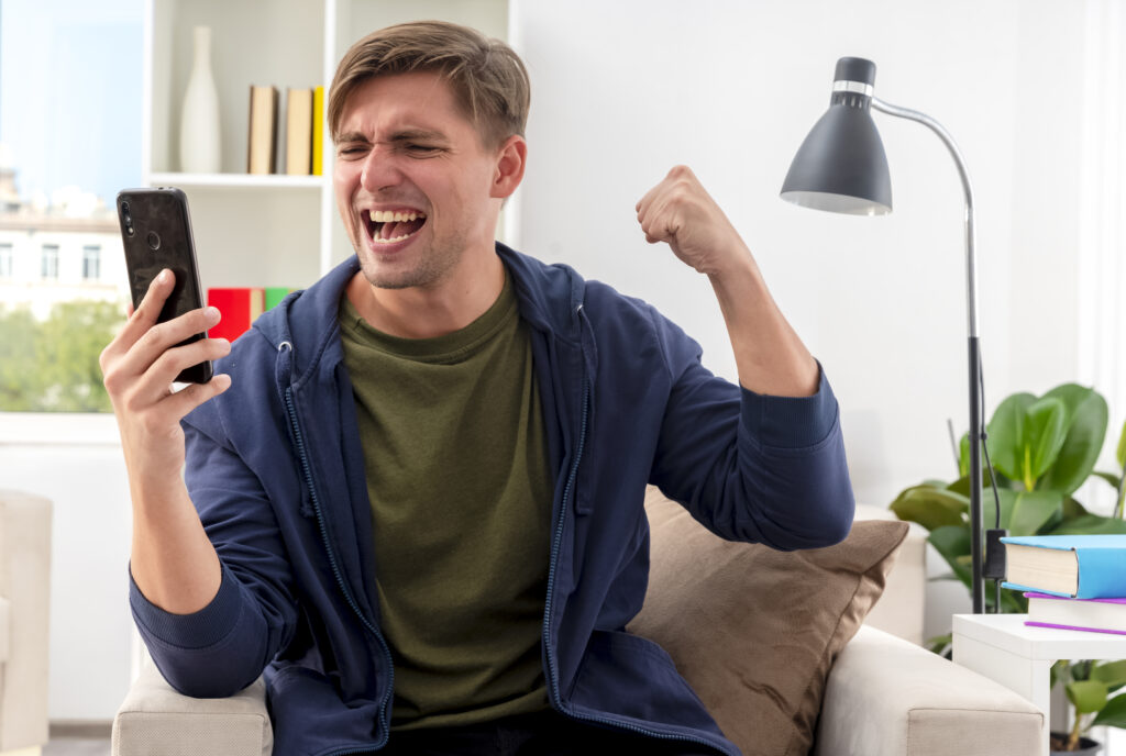 A young man sitting in an armchair, excitedly raising his fist in victory while looking at his phone, symbolizing success and achievement in the digital world.